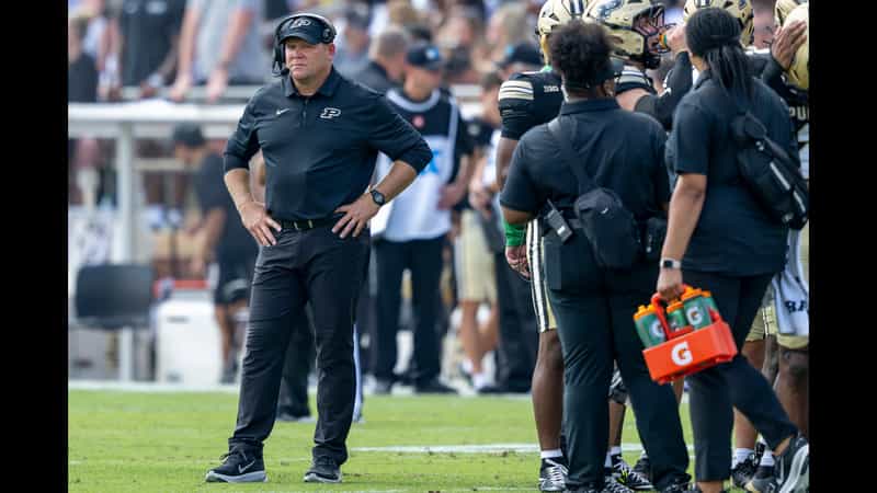 Purdue head coach Barry Odom stands on the sideline with hands on hips beside players and staff at Ross-Ade Stadium
