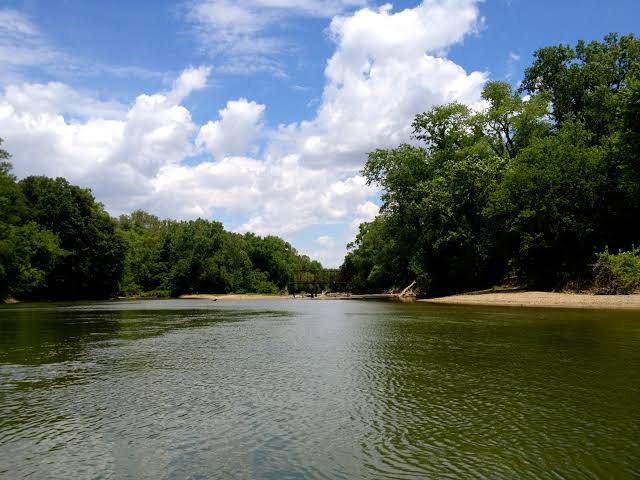 A wide, calm stretch of the Driftwood River in Indiana with a sandy bank on the right and green summer trees lining both sides under a partly cloudy sky.