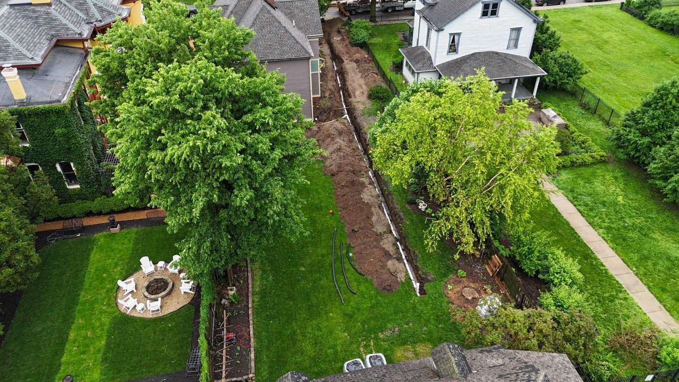 Aerial view of a long sewer line trench dug between two older homes in an Indianapolis neighborhood, with coiled black pipe staged on the lawn nearby