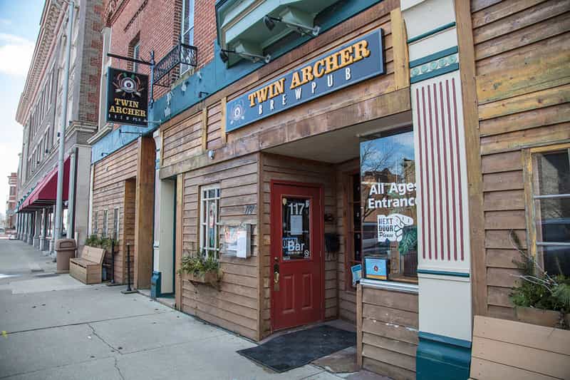 The wood-sided storefront of Twin Archer Brewpub on West Charles Street in downtown Muncie, with its brewpub sign and red All Ages Entrance door.