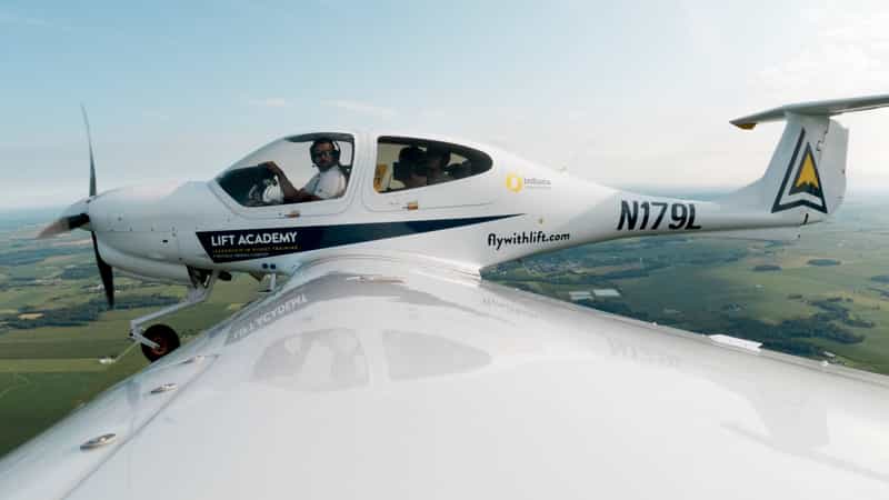 A LIFT Academy Diamond DA-40 training plane, tail number N179L, banks over green Indiana farmland with an instructor and student visible in the cockpit.