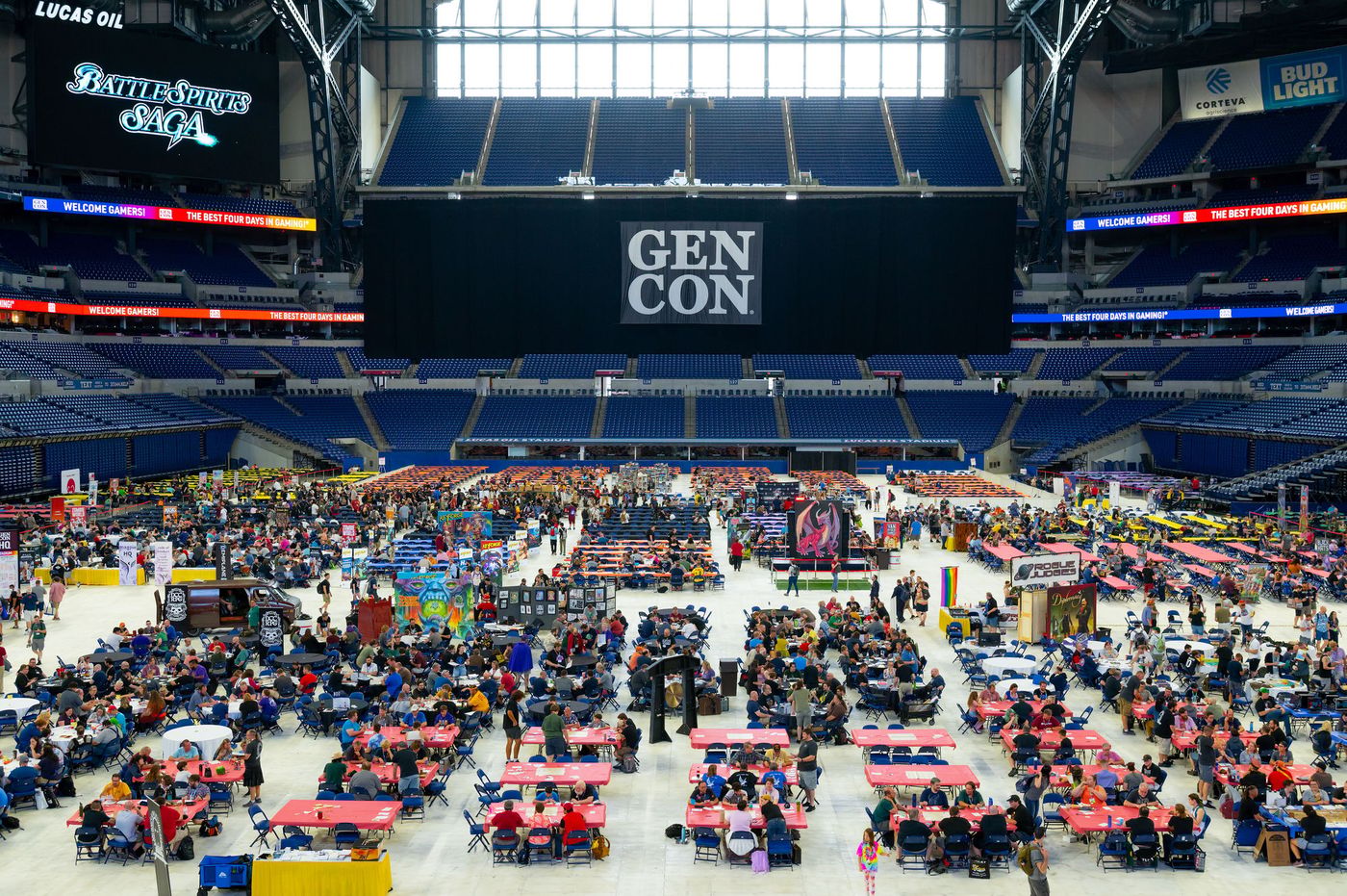 The Gen Con banner hanging above the gaming floor inside Lucas Oil Stadium in Indianapolis, with hundreds of gamers seated at long rows of tables