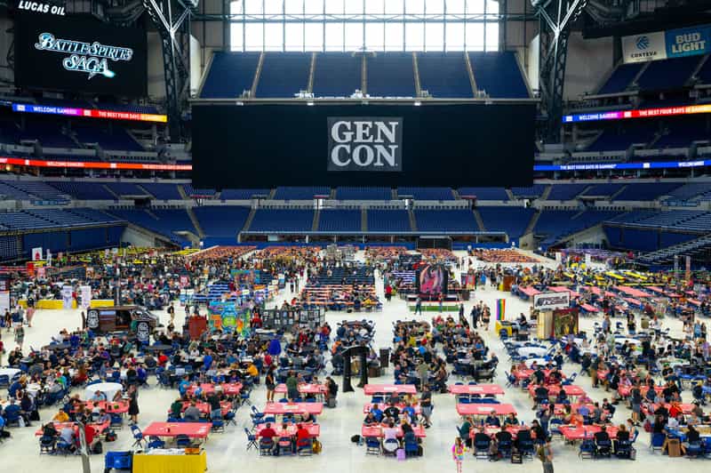 The Gen Con banner hanging above the gaming floor inside Lucas Oil Stadium in Indianapolis, with hundreds of gamers seated at long rows of tables