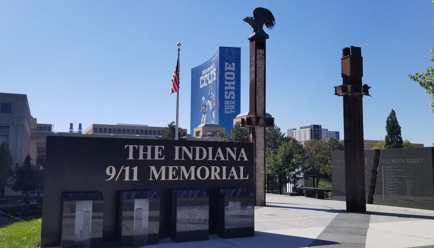 The Indiana 9/11 Memorial on the Downtown Canal in Indianapolis, with a black granite sign, two World Trade Center steel beams, a bronze eagle, and the Colts building in the background.