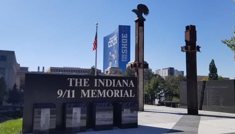The Indiana 9/11 Memorial on the Downtown Canal in Indianapolis, with a black granite sign, two World Trade Center steel beams, a bronze eagle, and the Colts building in the background.