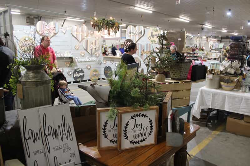 Indoor craft booths with rustic holiday signs ('Joy to the world', family signs), greenery and a shopper, inside the auction building