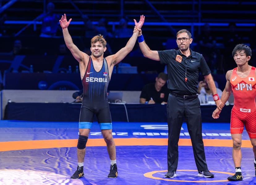 Stevan Mićić in a Serbia wrestling singlet raising both arms as the referee lifts his hand in victory at the 2023 World Championships in Belgrade