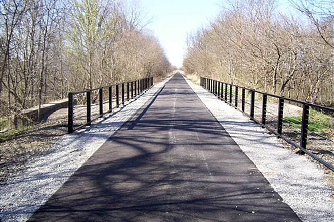 Paved Panhandle Pathway rail trail crossing a bridge with black metal railings, lined by bare early-spring trees