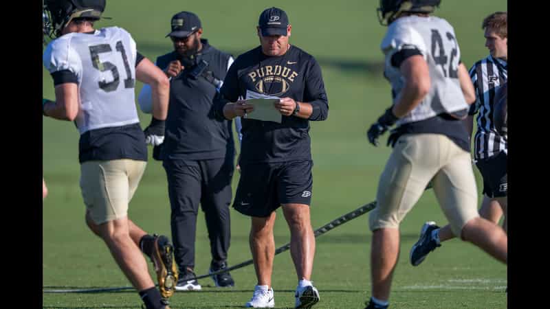 Purdue head coach Barry Odom walking the practice field with a call sheet as players in black and white jerseys run past during fall camp in West Lafayette
