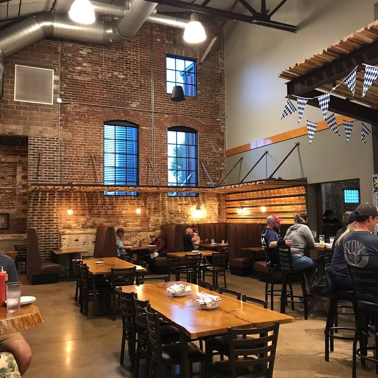 Diners at long wood tables beneath exposed brick walls, tall arched windows and blue-and-white Bavarian pennants inside Moontown Brewing Company's restored gymnasium in Whitestown, Indiana