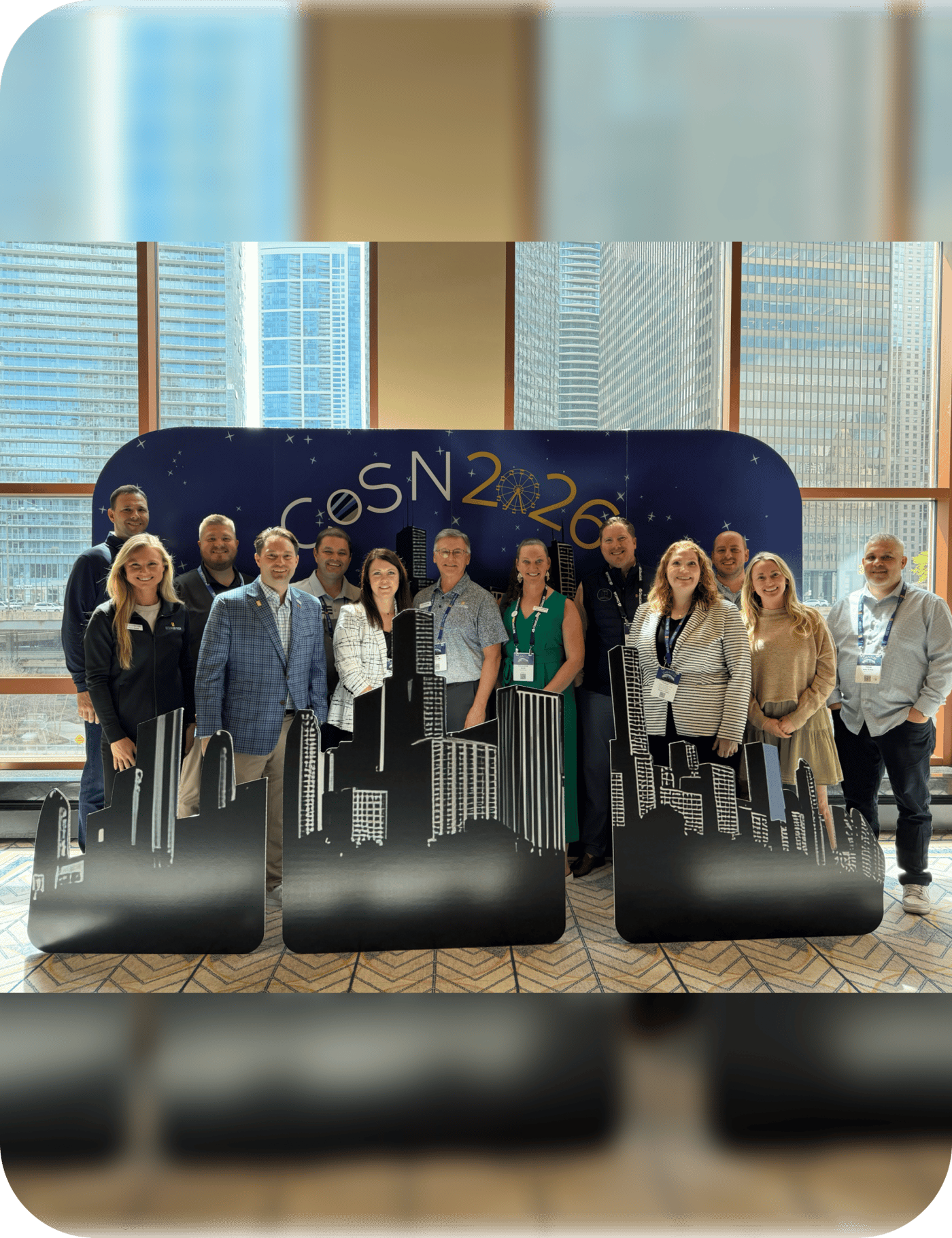 The Secured Tech team standing together in front of a CoSN 2026 conference backdrop and a Chicago skyline display, with the downtown Chicago skyline visible through the windows behind them.