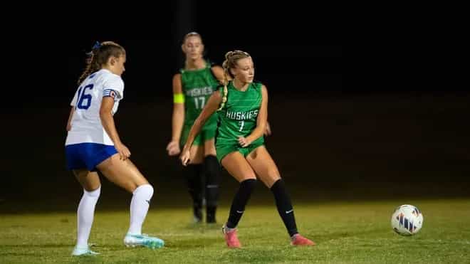 Evansville North player in a green Huskies jersey plays the ball as an Evansville Memorial defender in white closes in during a night girls soccer match