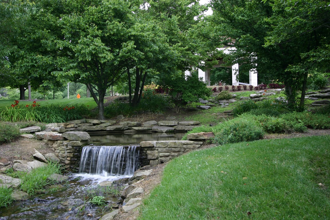 A stone-lined pond spilling over a small waterfall into a stream at Cox Arboretum in Bloomington, framed by shade trees, orange daylilies and a white columned pavilion on the rise behind