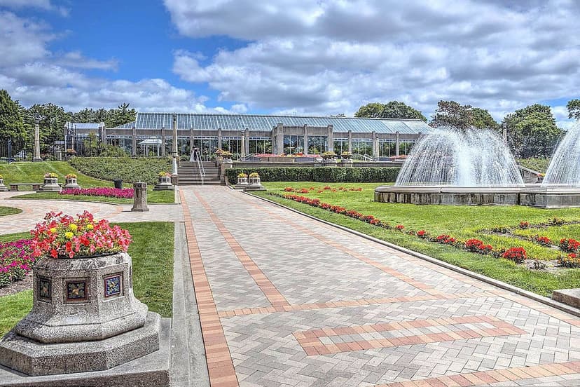 The Sunken Garden at Garfield Park in Indianapolis, with fountains running alongside a brick paver walkway and beds of red and pink flowers leading up to the glass conservatory building