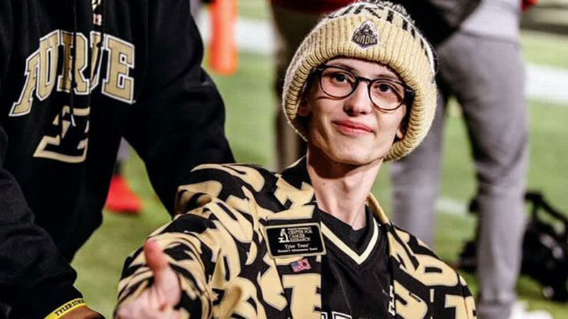 Tyler Trent smiling on the Ross-Ade Stadium sideline in a gold Purdue beanie and black-and-gold print jacket