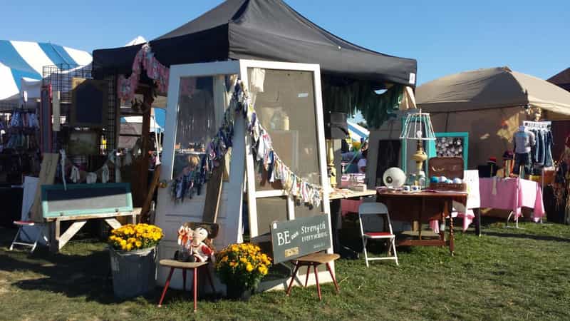Craft vendor booths on a lawn with a black canopy, vintage screen door, yellow mums and a "Be our strength" sign