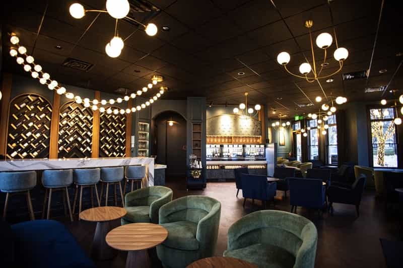 Dim wine bar interior with diagonal bottle racks, globe pendant lights, marble counter, bar stools and green armchairs