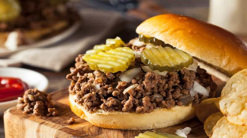 An Iowa loose-meat sandwich topped with dill pickle slices and diced onion on a bun, with potato chips on the side.