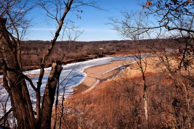 A view from a wooded bluff overlooking a partly frozen river and sandbar, framed by bare winter trees under a blue sky.