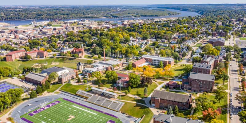 Aerial view of a tree-lined Iowa college campus with a football field, the surrounding town, and a river in the distance.