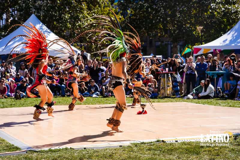 Performers in feathered regalia dance before a seated crowd at an outdoor Latino heritage festival in Iowa.