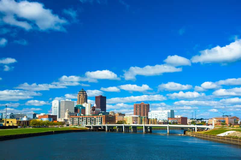 The downtown Des Moines skyline across the Des Moines River and a bridge, under a bright, partly cloudy blue sky.
