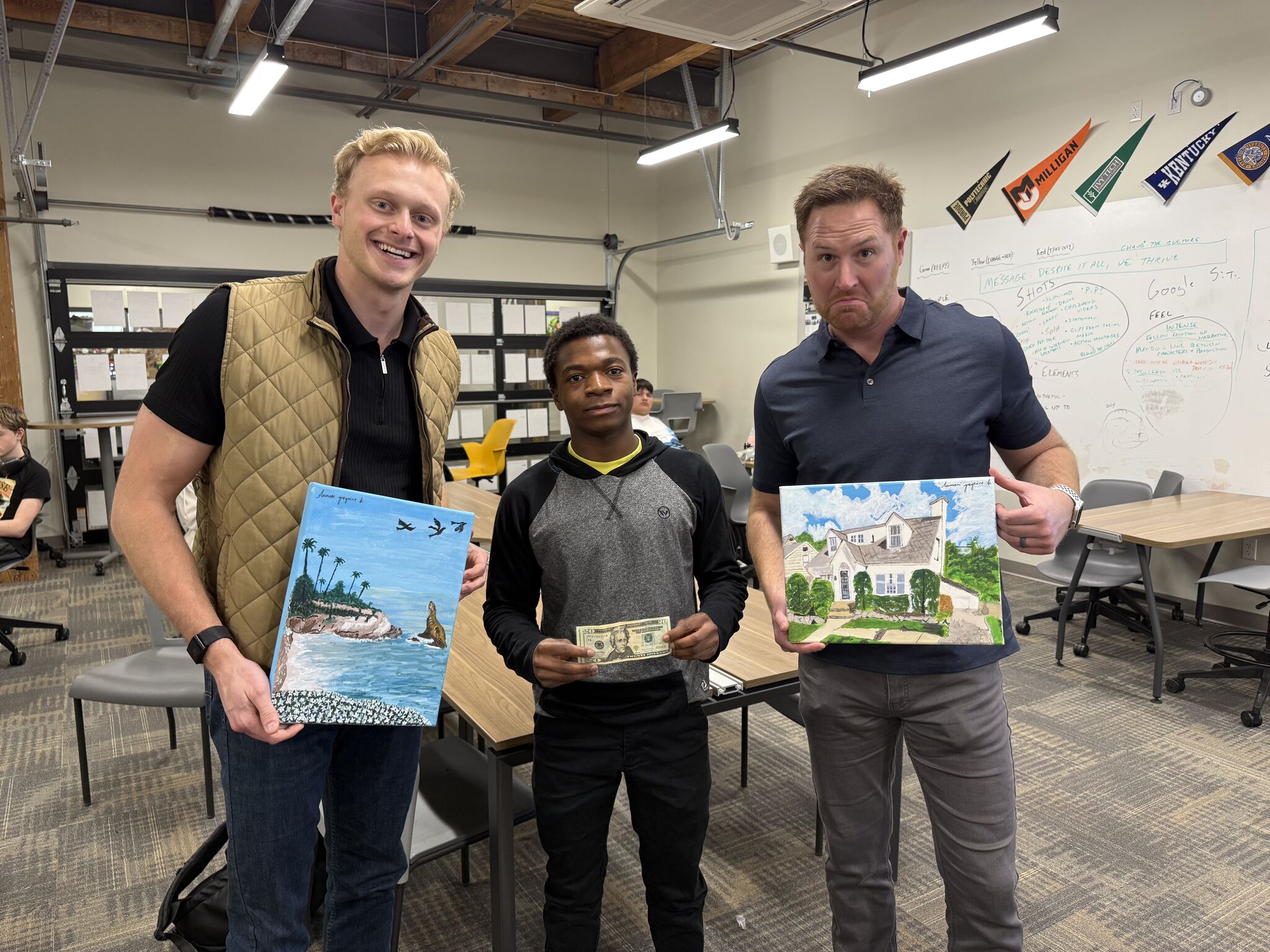 A student holding his cash earnings in a classroom, flanked by two mentors holding framed prints.
