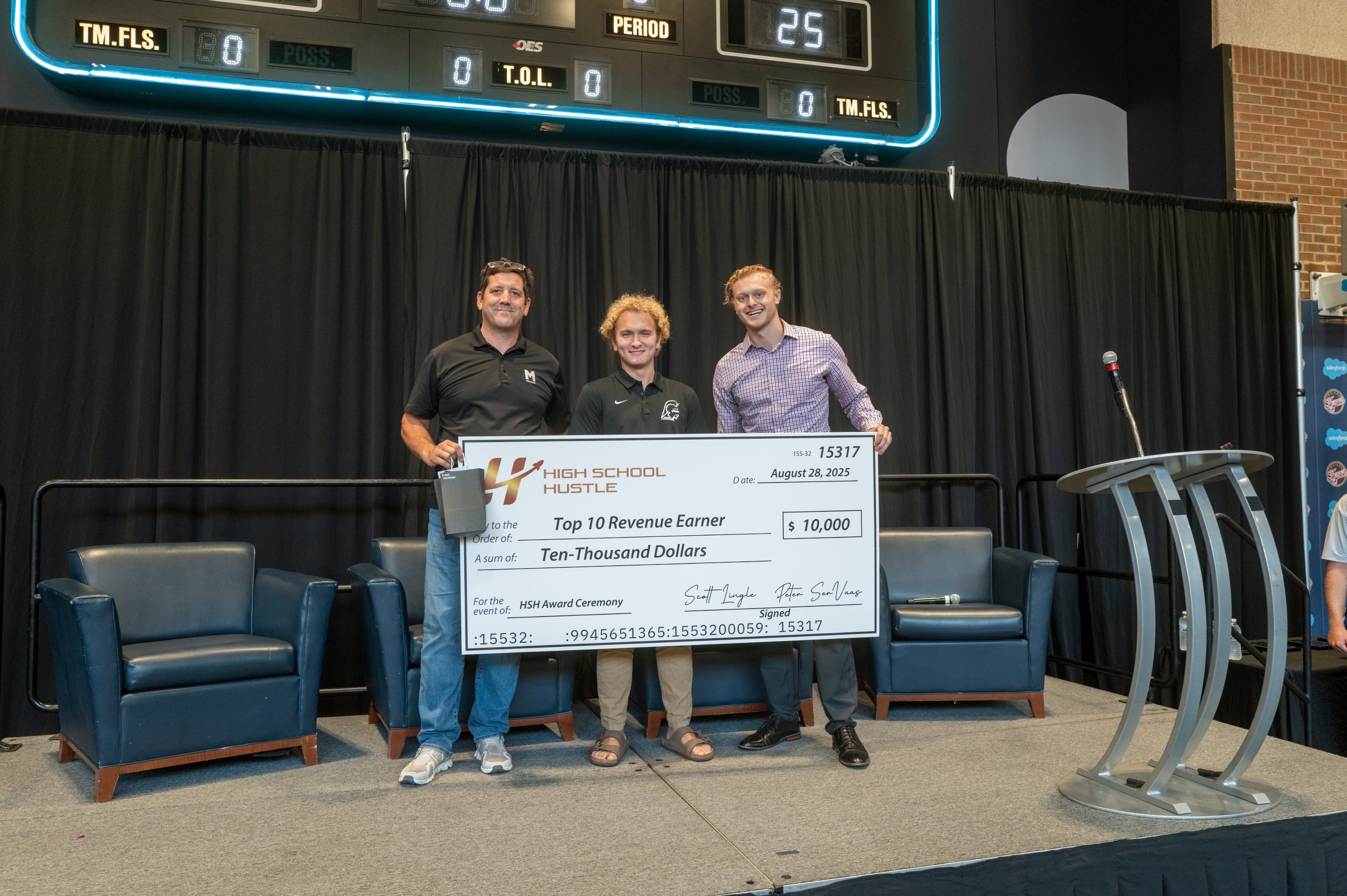 High School Hustle award winners on stage holding an oversized check under an arena scoreboard.
