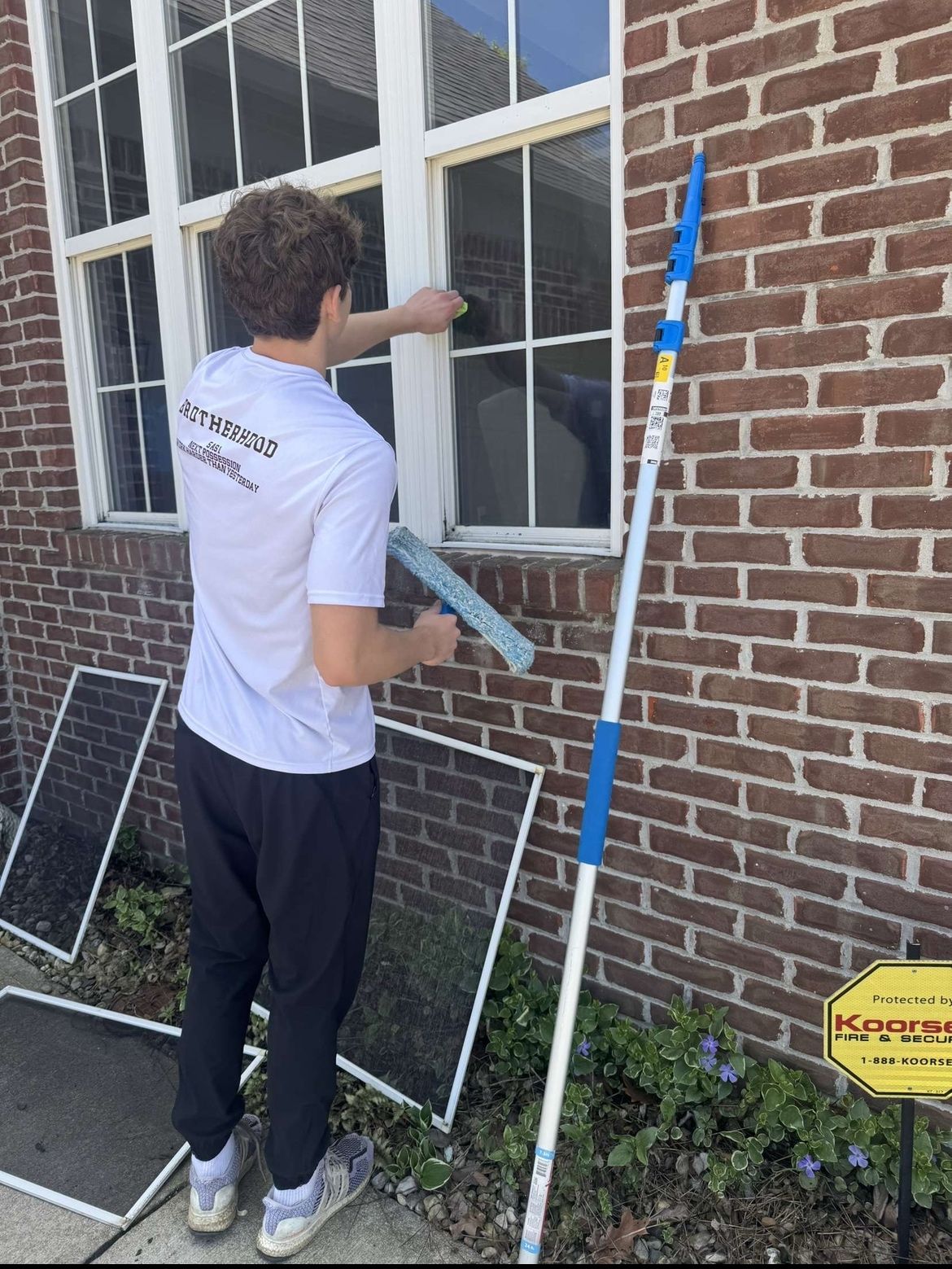 A student cleaning a window on a brick house with a squeegee and an extension pole.