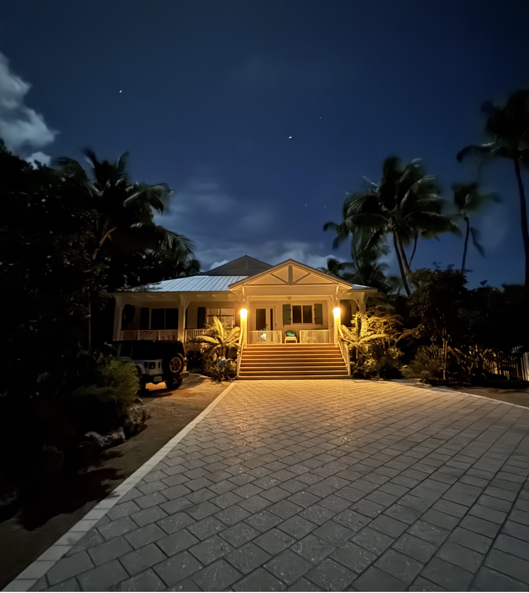 A Florida Keys house at night, warm lights glowing on the front porch at the top of a paver driveway, palm trees and stars overhead.