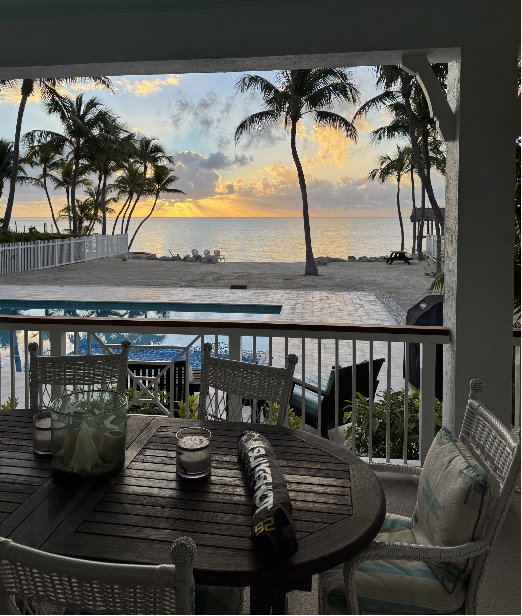 Sunset over the water seen from a covered porch in the Florida Keys, with palm trees, a pool and a wooden table in the foreground.
