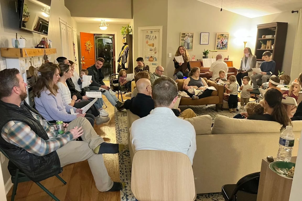A District Church gathering in a living room, singing together from song sheets with children present
