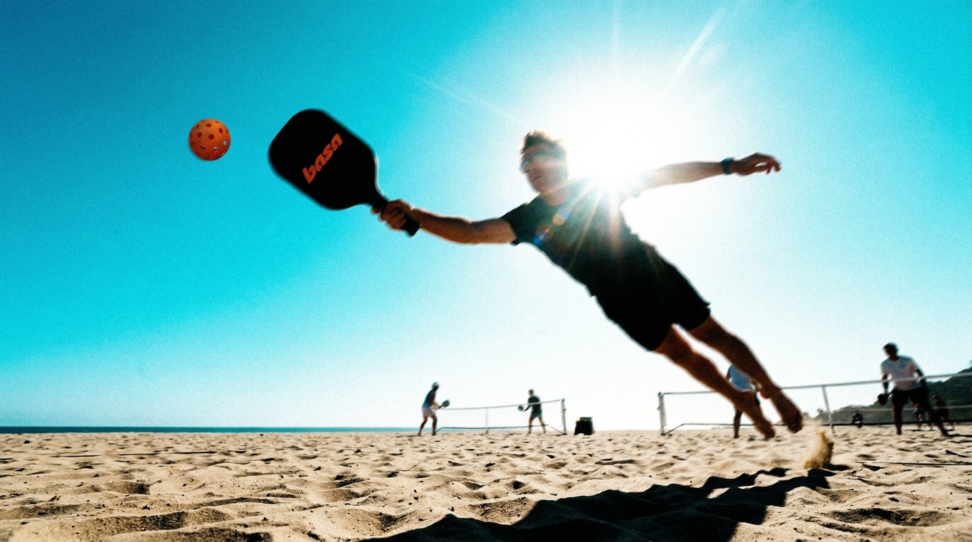 Player diving mid-air to strike a Basa pickleball on a sun-drenched beach court