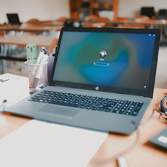 Laptop 14 cali z otwartym dziennikiem elektronicznym na biurku nauczyciela, obok stos zeszytów i kubek