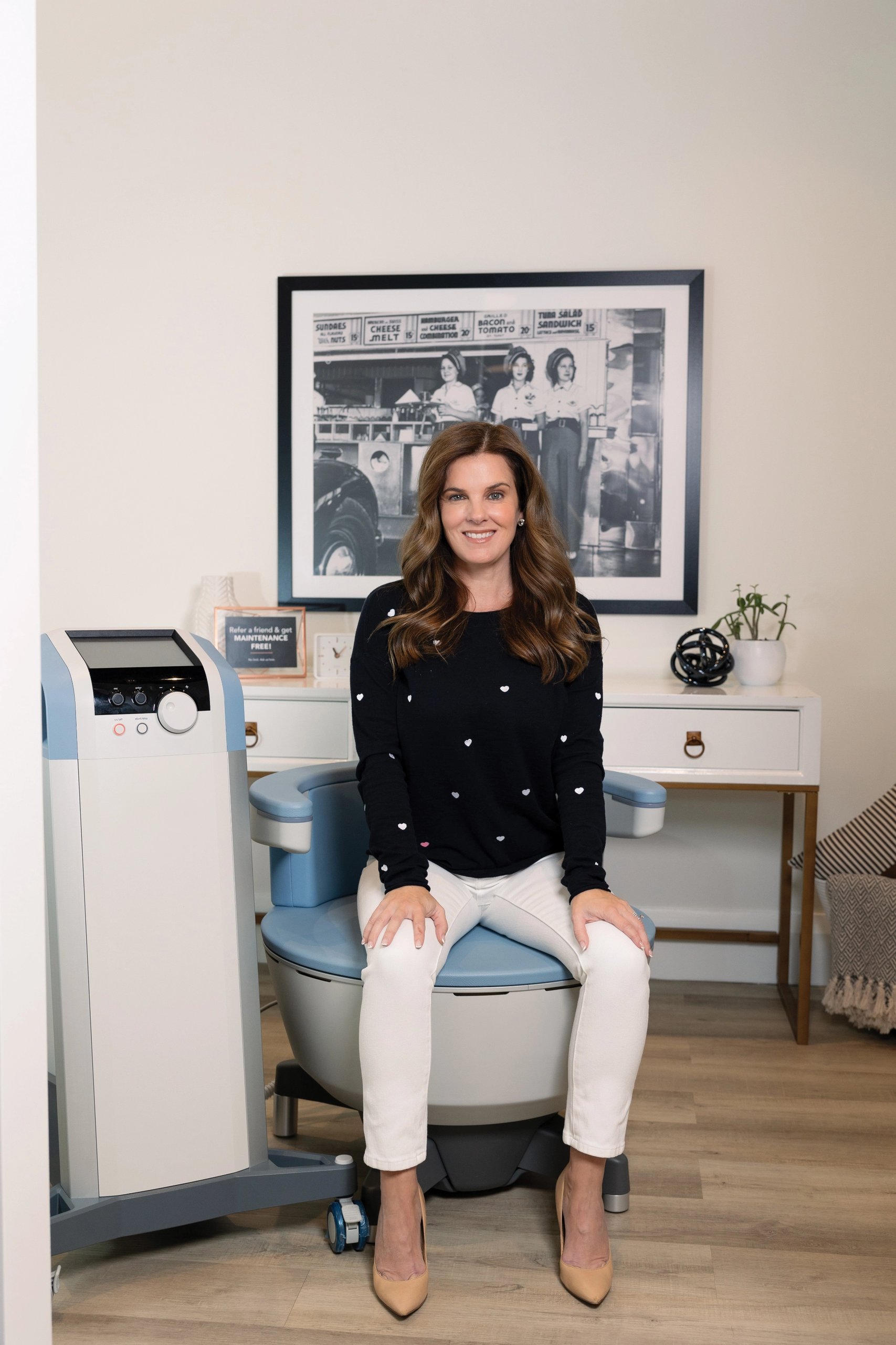 Woman sitting next to a UROSPOT® device in a modern interior setting, wearing a black turtleneck and beige skirt.