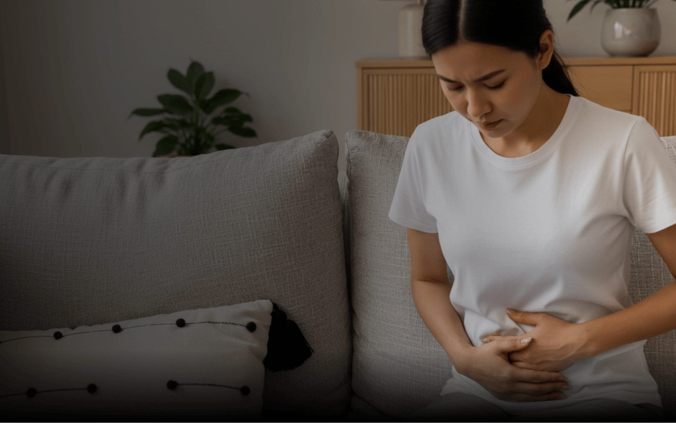 A woman in a white shirt sits on a sofa, holding her lower abdomen while seeking pelvic organ prolapse help in Halifax.