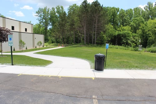 Hole 1 starts off the parking lot. A set of pine trees guard the entrance to the green. Try not to throw on top of the gym! (Practice basket featured on right)