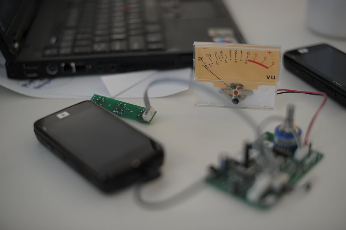Analog VU meter on a table beside a Nokia N900, green circuit boards, connecting cables, and a laptop.