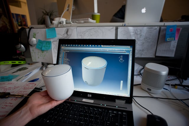 A hand holds a Social Speaker shell in front of a laptop displaying a three-dimensional model.