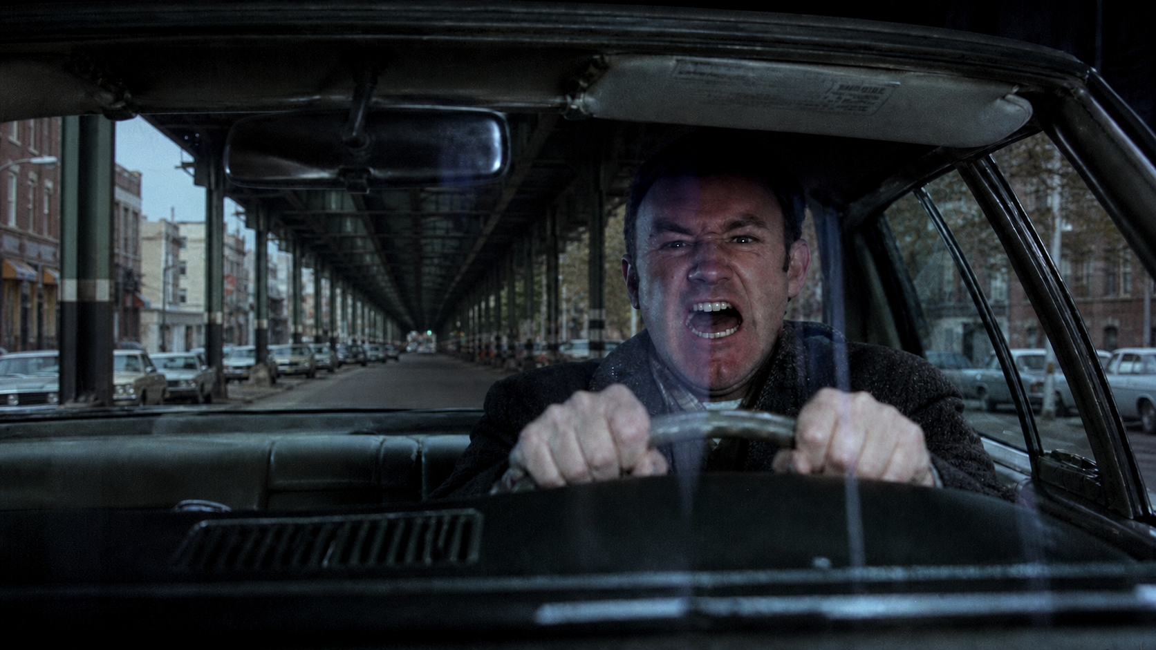 The French Connection film still with a man driving directly beneath a green New York City elevated train line in Queens.