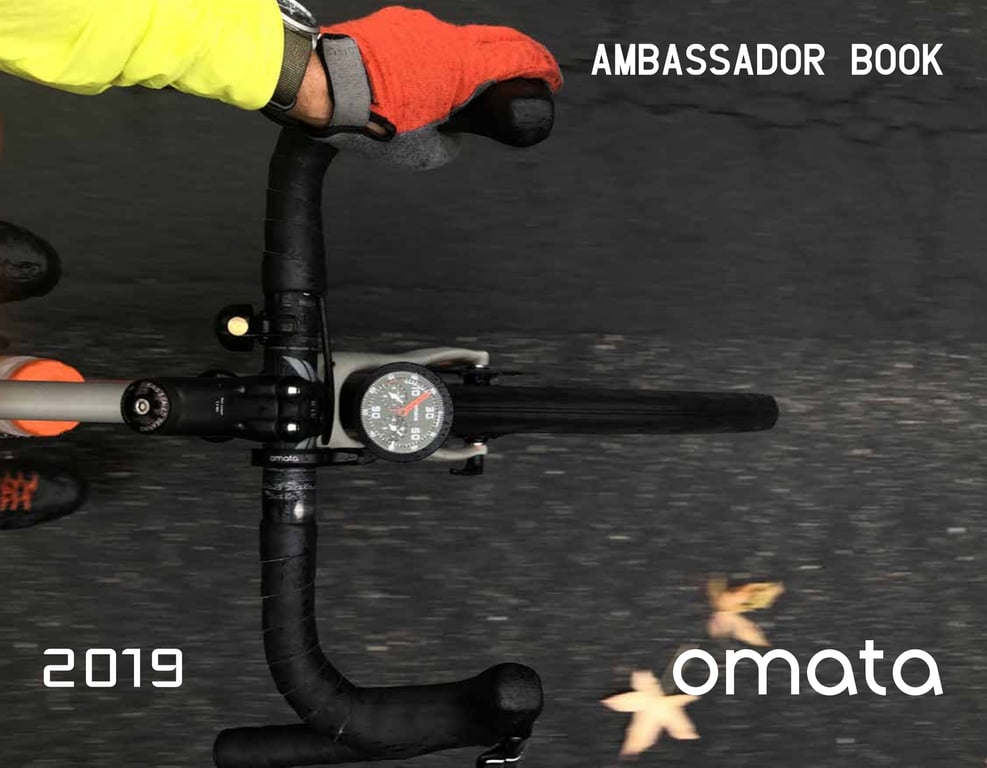 March 2019 OMATA Ambassador Book cover showing a cyclist's orange glove on handlebars with the OMATA One against a dark road.