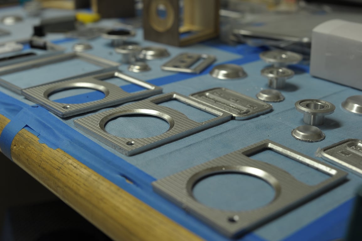 Metal Humbo faceplates with circular speaker openings and rectangular meter openings lie beside machined control fittings on a blue-covered bench.