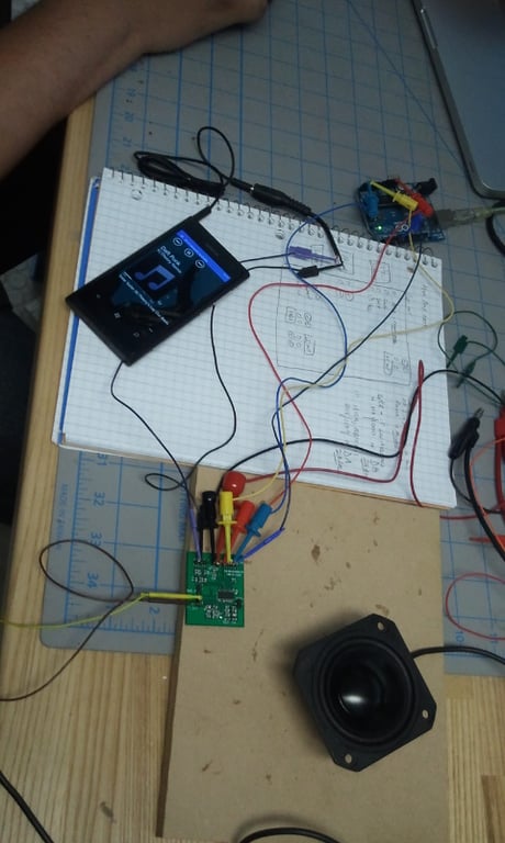 Top-down view of a phone, prototype circuit boards, a speaker driver, cables, and sketches during Social Speaker electronics testing.