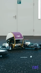 A small robot with exposed wiring moves on a speckled floor near a door.
