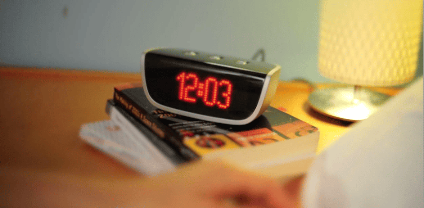 A silver and black Social Alarm Clock showing 12:03 in red numerals sits on a stack of books beside a lit bedside lamp.