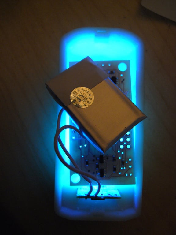 Open Kombolói enclosure glowing blue around a circuit board, battery, and looped wires.