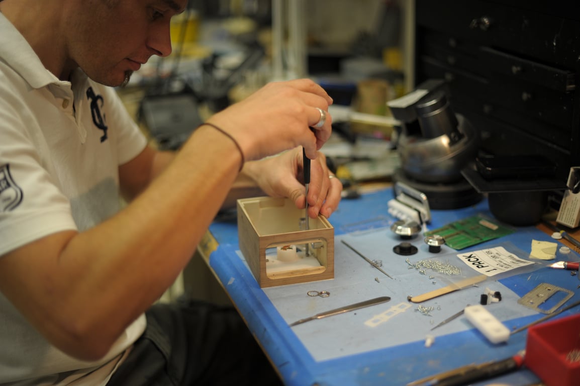 A person uses a screwdriver inside a wooden Humbo enclosure at a workbench covered with tools and metal fittings.