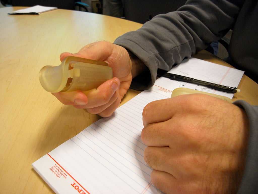 A hand holding a translucent enclosure with internal mounting features above a table and lined notebook.