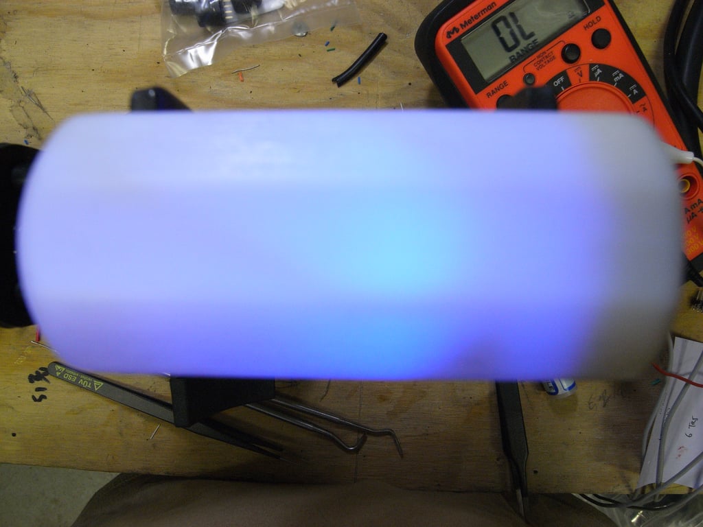 White Kombolói enclosure glowing blue on a wooden workbench beside an orange multimeter.