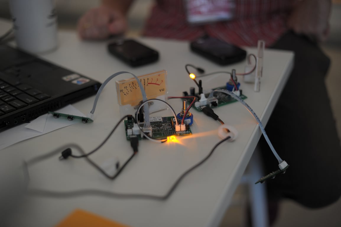 Two connected circuit-board assemblies with glowing indicator lights, an analog meter, cables, phones, and a laptop on a table.